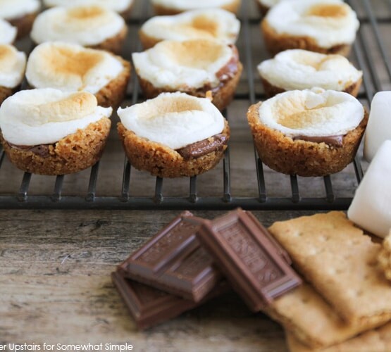 several mini smores bites fresh from the oven sitting on a cooling rack. a few pieces of chocolate, marshmallows and graham crackers are piled in front of the rack.