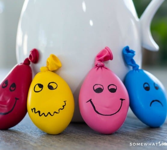 a red, yellow, pink and blue homemade stress ball leaning up against a pot on a table. Each DIY stress ball has a funny face drawn on each one.