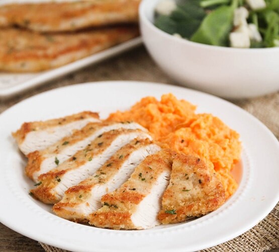 sliced Turkey Cutlets with a side of mashed potatoes on a white plate with a bowl of salad in the background