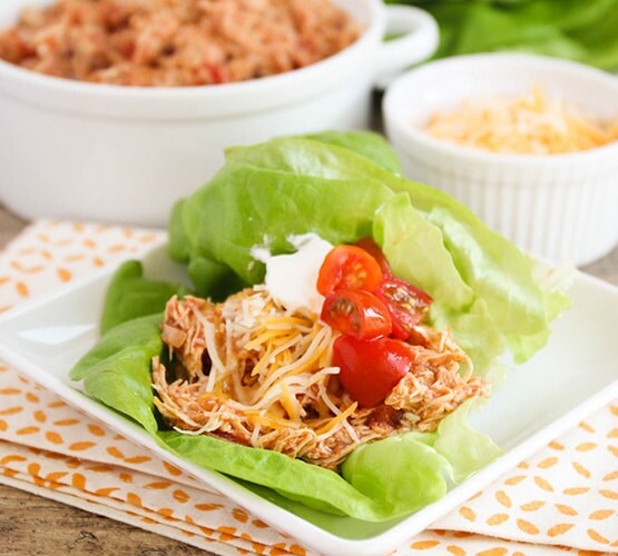 a square white plate with two Chicken Taco Lettuce Wraps on it. Each wrap is topped with shredded cheese cherry tomatoes. Behind the plate is a bowl of shredded chicken and a smaller bowl of shredded cheese.