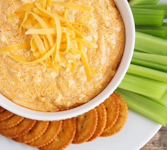 looking down on a white bowl full of this easy buffalo chicken dip recipe that was made in a crock pot and topped with shredded cheese. On the plate next to the bowl are several Ritz crackers and celery sticks