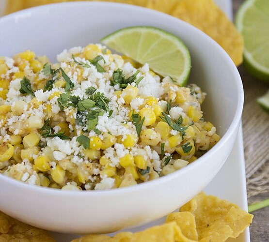 a bowl of Mexican corn salad topped with cilantro and cotija cheese on a serving dish next to tortilla chips