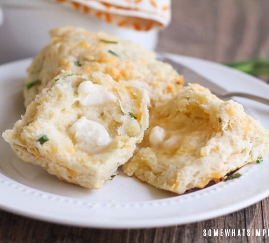 two cheddar biscuits with chives on a plate. One is cut in half with a pad of melted butter on top.