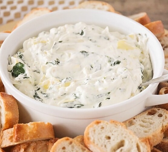 a white bowl filled with Cheesy Spinach Artichoke Dip that has a spoon stuck in the dip. Surrounding the bowl are slices of baguettes.