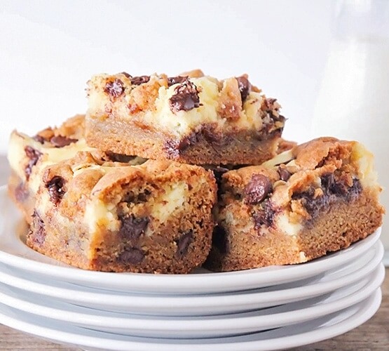 a pile of cheesecake cookie bars on a stack of white plates. Next to the plates are a glass pitcher of mile and a red and white striped straw laying down.