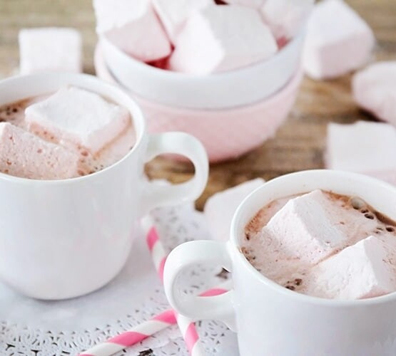 two cups of hot chocolate that have Homemade Strawberry Marshmallows floating in them. Behind the mugs is a bowl of more marshmallows.