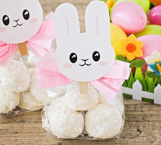 two packages of Easter bunny tail cookies wrapped in plastic with a pink bow and topped with an Easter bunny printable. Next to the cookie gift bags is an Easter basket filled with plastic eggs.