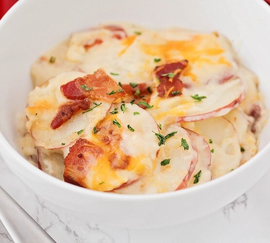 looking down on a white bowl filled about halfway with scalloped potatoes and topped with cheddar cheese, bacon and parsley. A fork is next to the bowl and a red checkered cloth napkin is laying behind the bowl.