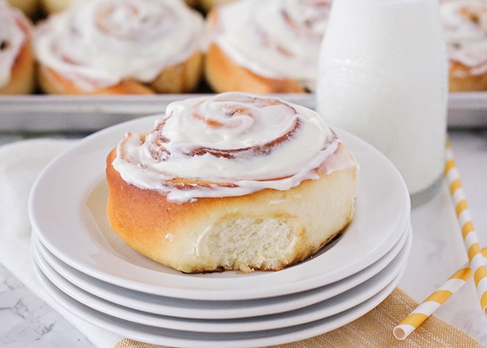a homemade Cinnamon Roll topped with a cream cheese frosting on a stack of white plates with a jar of milk and a tray of cinnamon rolls in the background.