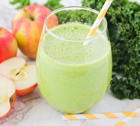 a glass filled with a green smoothie and a yellow striped straw in the cup. Next to the glass on the counter are apples, leaves of kale and 2 more striped yellow straws.