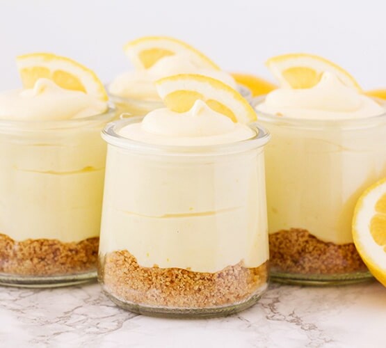 four cups of lemon cheesecake mousse topped with lemon slices and lemons next to the cups on the counter