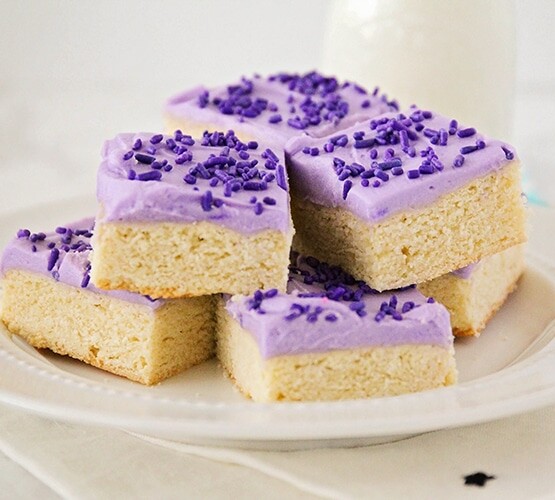 a white plate full of sugar cookie bars that are stacked on top of each other. Each cookie bar is topped with purple frosting and purple sprinkles. A glass of milk is on the counter behind the plate of sugar cookie bars.