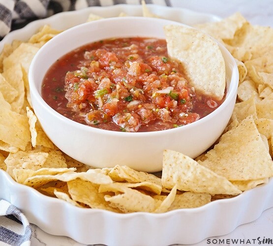 a white bowl filled with this easy blender salsa recipe with a tortilla chip standing inside the bowl. This bowl is in the center of a white serving dish with tortilla chips all around the bowl.