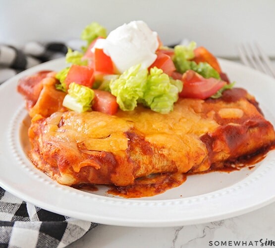 two enchilada style baked bean burritos on a white plate topped with lettuce, tomatoes and sour cream. Next to the plate is a black and white checkered cloth napkin.