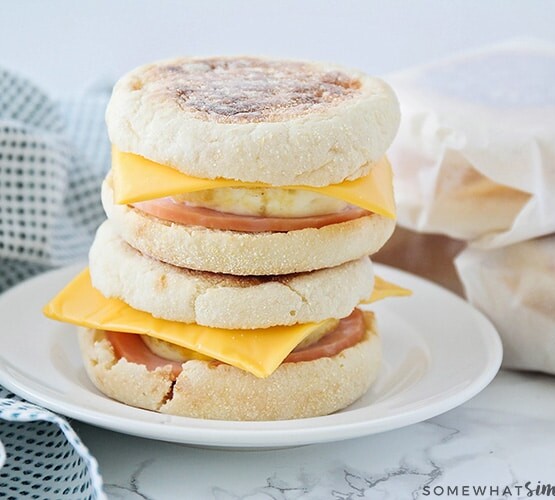 Two make ahead breakfast sandwiches stacked on top of each other on a white plate. Each one is made with an English muffing, American cheese, eggs, and Canadian bacon. Behind the plate are three more sandwiches wrapped in parchment paper.