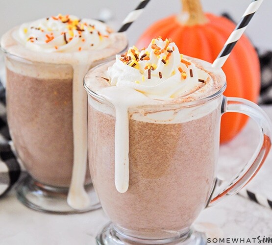 two glass mugs filled with a homemade pumpkin spice hot chocolate topped with whipped cream and sprinkles