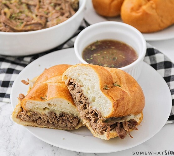 a french dip sandwich cut in half and stacked on a white plate with a side of au jus in a small white bowl. Behind the plate is a bowl filled with shredded beef and a plate with French rolls on it.