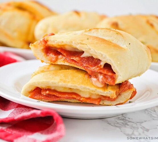 a homemade pepperoni pizza pocket that has been cut in half and the two halves are stacked on top of each other. In the background is a tray filled with several more golden brown pizza pockets.