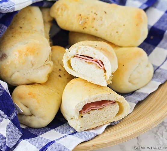 a bowl of baked Brazilian ham and cheese rolls that are golden brown and wrapped in a blue and white checkered cloth napkin. One of the rolls has been cut in half and you can see the smoked ham and mozzarella cheese inside.