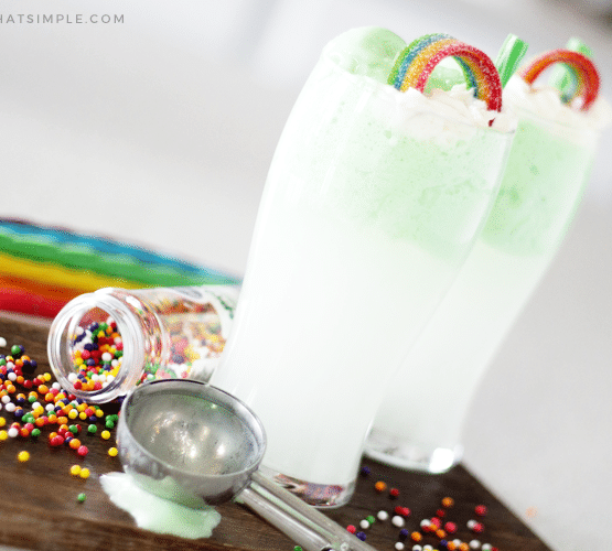 two green st. patrick's day drinks on a cutting board topped with a rainbow candy and a green piece of licorice for a straw. There are colored sprinkles spilled on the cutting board with an ice cream scoop laying next to the drink and colored pieces of licorice in the background.