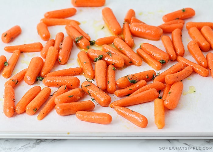 a bunch of baby carrots have been spread over a large baking sheet covered with parchment paper. The carrots have been coated in olive oil and honey and ready to be baked in the oven