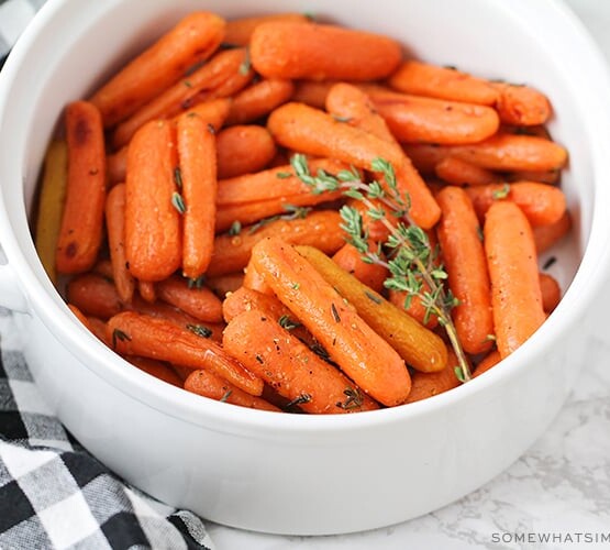 a white serving bowl that is filled with honey glazed carrots that have been baked and topped with thyme. A black and white checkered napkin is laying next to the bowl on the counter.