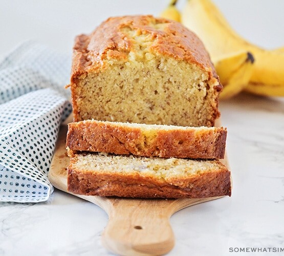 a loaf of homemade banana bread made with this easy recipe . Two slices have been cut off from one end and are laying down on the cutting board. Behind the loaf of bread is a small bunch of bananas.