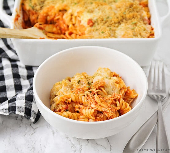 a white bowl filled with a serving of chicken parmesan casserole that is made with pasta and topped with bread crumbs and cheese. Behind the bowl on the counter is a casserole pan filled with the remaining casserole.