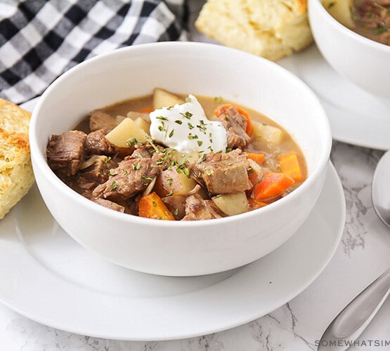 a white bowl filled with savory crock pot beef stew. There are chunks of beef and chopped potatoes and carrots topped with a dollop of sour cream and chopped parsley. The bowl is sitting on a white plate with a square of bread next to it. Above the bowl is a black and white checkered cloth napkin and in the corner of the image another bowl of stew is partially visible.