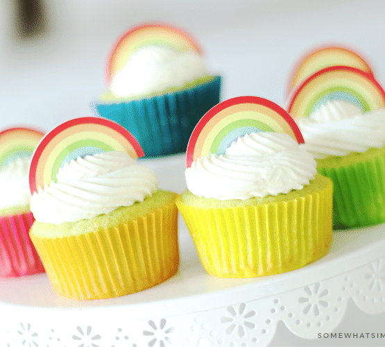 a close up of Key Lime Cupcakes sitting on top of a white cake stand. The lime cupcakes are wrapped in different colored cupcake wrappers and topped with white frosting and a rainbow cupcake topper