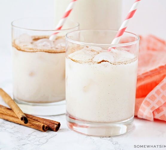 two glasses filled with this easy horchata recipe. Each glass has a red and white striped straw in it. Next to the glasses are 4 cinnamon sticks laying on the counter as well as a light red cloth napkin.