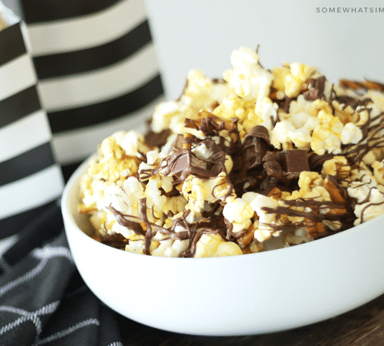 a white bowl filled with gourmet popcorn with drizzled chocolate on top mixed with pieces of kit kat bars and pretzels. Behind the bowl are two black and white striped bags filled with more chocolate covered popcorn