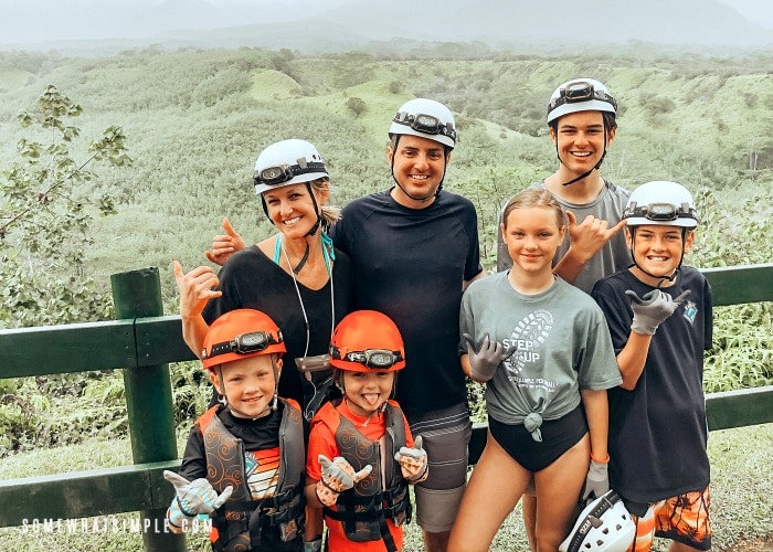 an attractive mom and dad with their five kids most wearing helmets with head lamps, who are getting ready for a tubing adventure, overlooking lush green hills in kauai