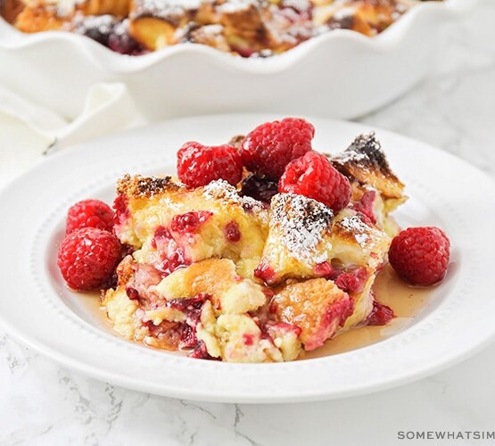 a white plate filled with baked raspberry french toast that is topped with fresh whole raspberries and syrup