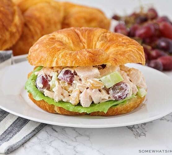 a chicken salad sandwich made with a croissant on a white plate. More croissants and a pile of grapes are laying on the counter behind the plate