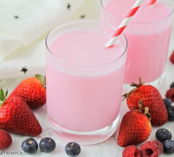 a glass filled with a pink yogurt drink with a striped straw in the cup. Surrounding the glass are several strawberries, blueberries and raspberries.