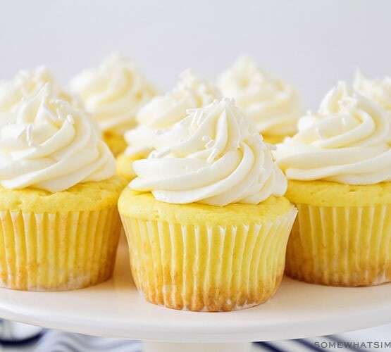 a white cake stand with several lemon cupcakes topped with a cream cheese frosting and white sprinkles. There are a couple of lemons on the counter next to the cake stand.