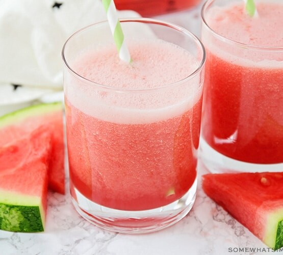 two glasses filled with this watermelon cooler recipe. Each glass has a green and white striped straw and slices of watermelon are next to the cups on the counter