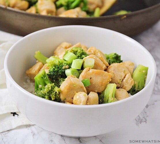 a white bowl filled with broccoli and chicken stir fry topped with chopped green onions all over a bed of white rice