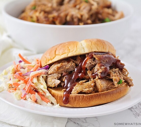 a pulled pork sandwich made in a slow cooker topped with bbq sauce and a side of coleslaw on a white plate.