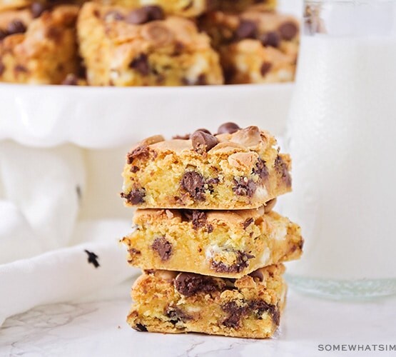 a stack of chocolate chip cookie bar squares made from cake mix next to a jar of milk with a cake stand filled with more bars behind it.