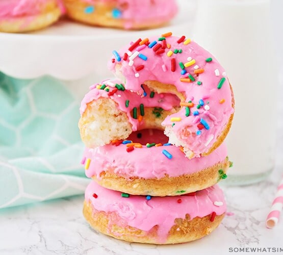 a stack of cake mix donuts, topped with pink icing and colored sprinkles. The donut on top has been cut in half.