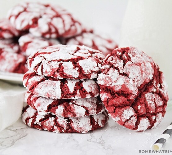 a stack of red velvet cake mix cookies with a dusting of powdered sugar next to a glass jar of milk. Behind these are another plate full of more cookies.