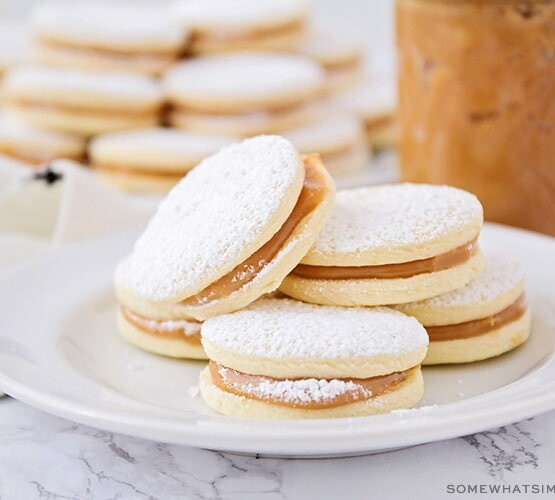 a plate of homemade alfajores with powdered sugar on a table made using this easy recipe with a jar of dulce de leche behind it