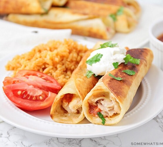 two crispy chicken taquitos on a plate topped with sour cream and a side of rice