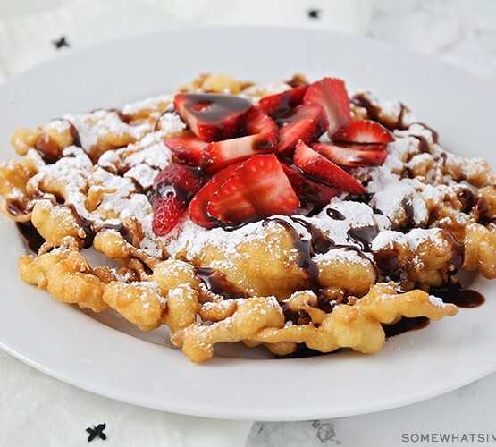 a homemade funnel cake on a white plate topped with freshly sliced strawberries, powdered sugar and chocolate syrup