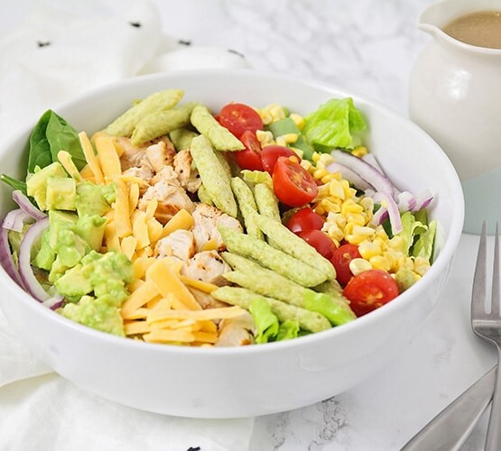 grilled chicken salad with lettuce, cheese, corn, tomatoes, and snap peas in a white bowl